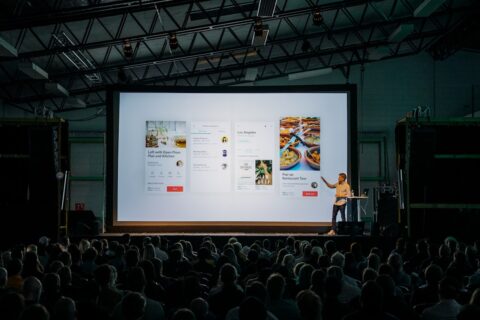 person discussing while standing in front of a large screen in front of people inside dim-lighted room