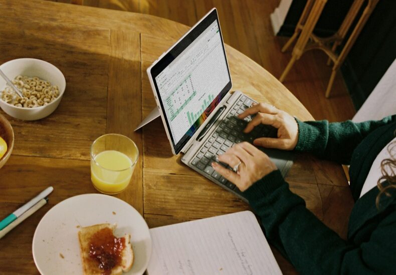 Person working on laptop at wooden table with breakfast.