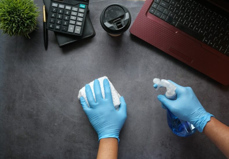 Hands in blue gloves cleaning a dark desk with a spray bottle and cloth